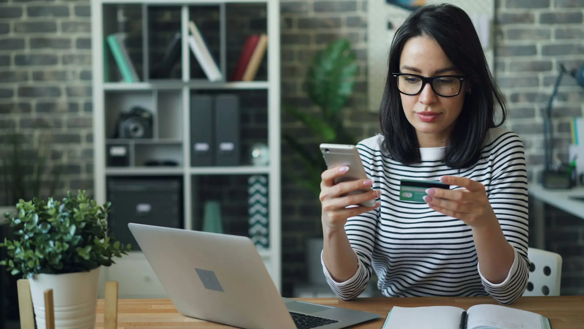 Woman holding a bank card while using her phone at a laptop